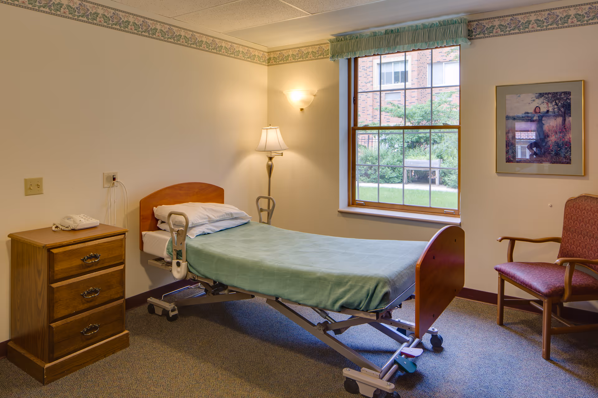 A small, simple bedroom in a senior living facility with a single hospital-style bed covered with a green blanket. Next to the bed is a wooden nightstand with a telephone on top. A floor lamp and a wall-mounted light provide illumination. There is a large window with a view of greenery outside, and a framed picture hangs on the wall above a red upholstered chair.