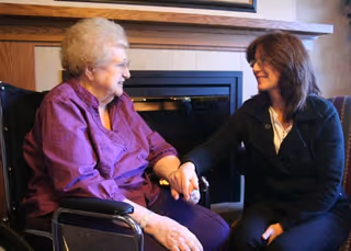 An elderly woman sitting in a wheelchair holding hands and smiling with a younger woman seated next to her in a cozy room with a fireplace in the background.