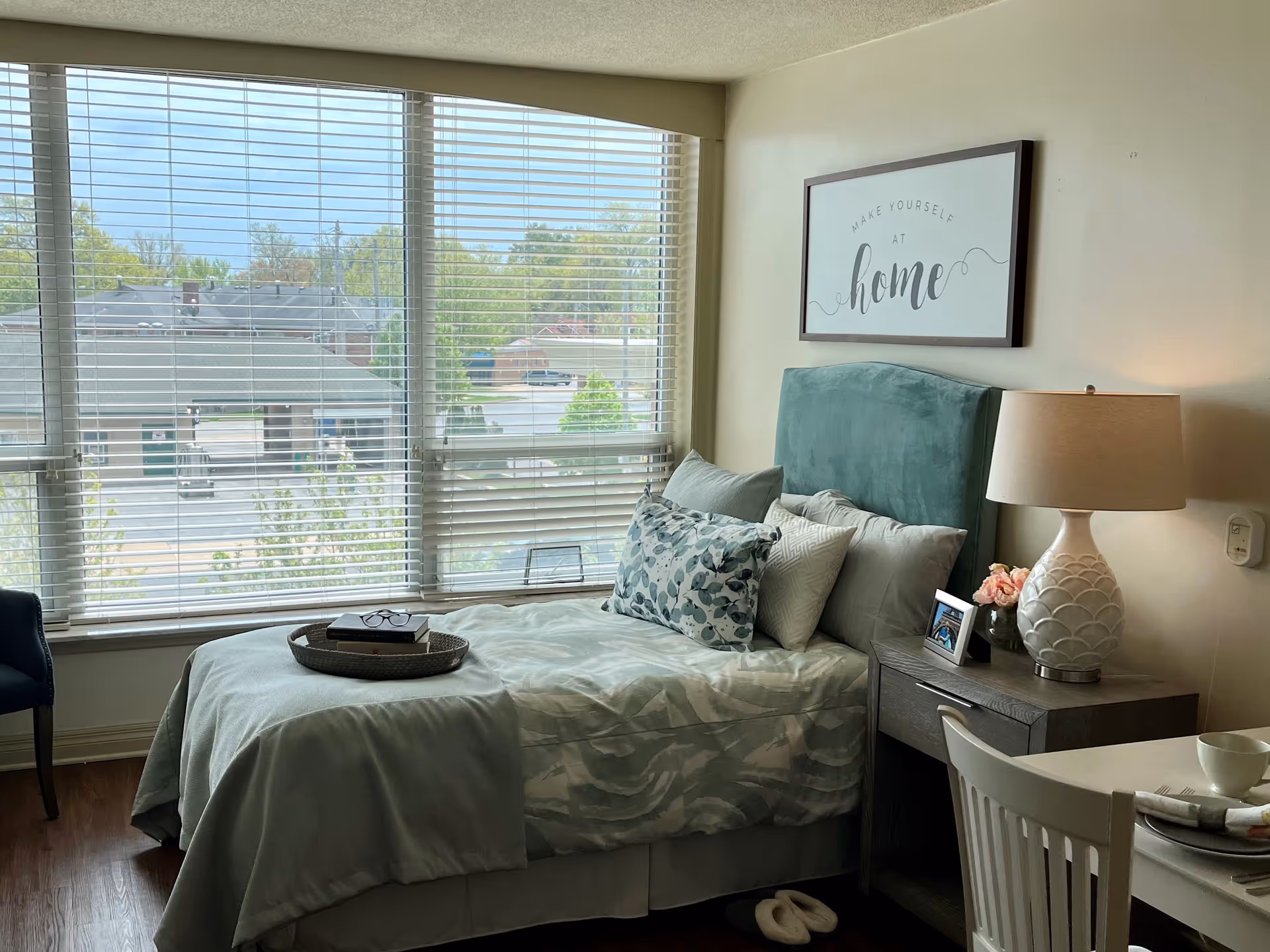 A cozy bedroom with a large window covered by blinds, a bed with multiple pillows and a teal headboard, a nightstand with a lamp, a small vase with flowers, and a framed photo. A framed sign above the bed reads 'Make yourself at home'. There is a chair and a table with a cup and utensils in the foreground.