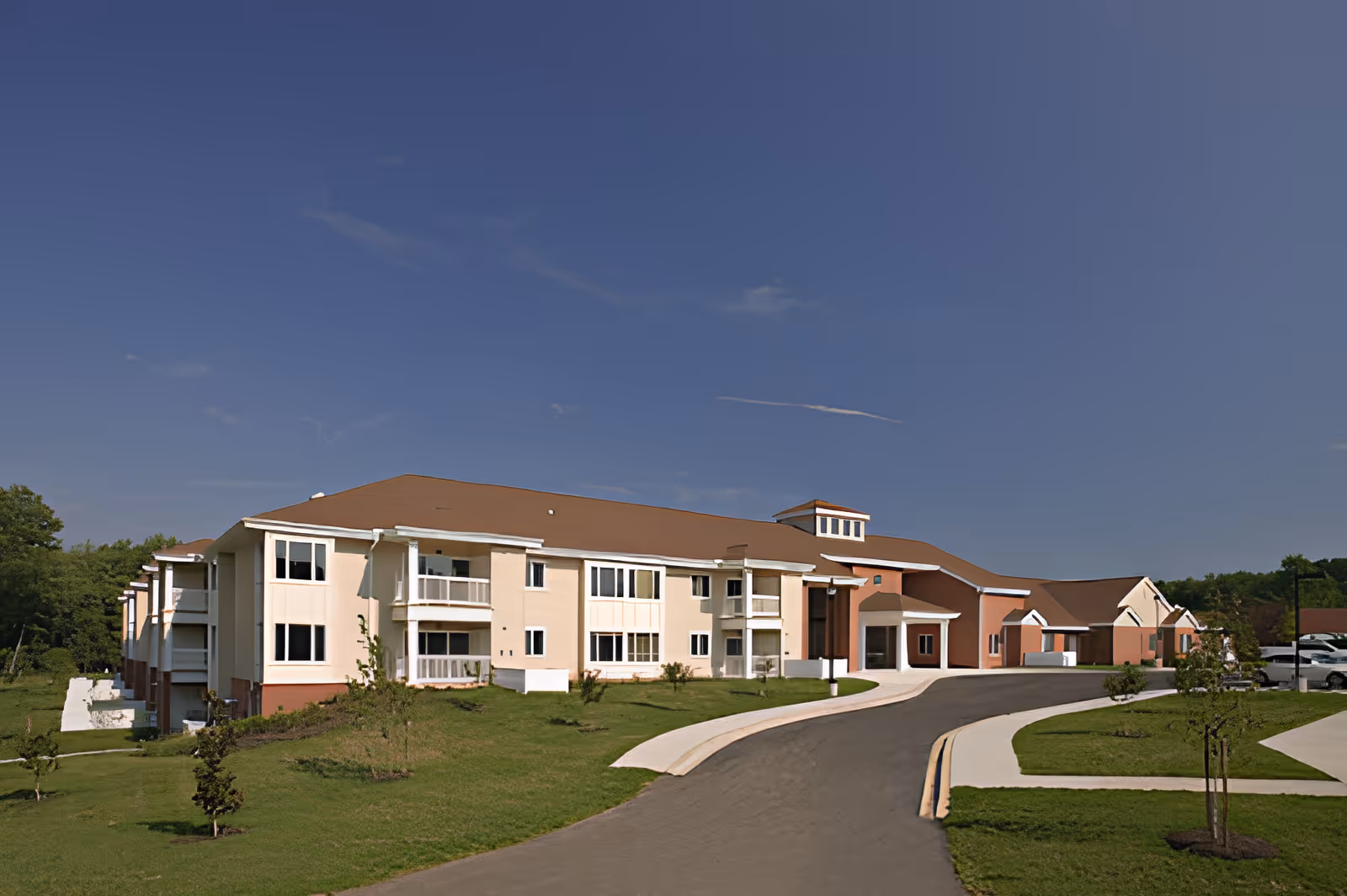 Exterior view of a large, two-story senior living facility building with beige and brown walls, multiple windows, balconies, and a curved driveway leading to the entrance. The building is surrounded by a well-maintained lawn with small trees and shrubs under a clear blue sky.