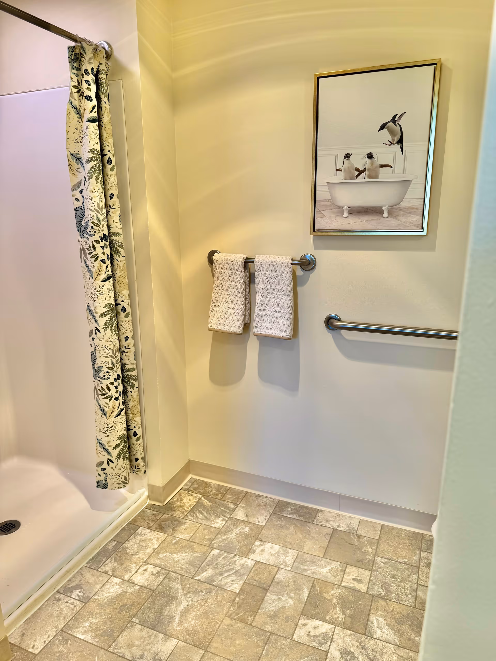 Corner of a bathroom showing a shower with a patterned curtain, towel bars holding two towels, a grab bar, tiled floor, and penguin bathtub wall art.