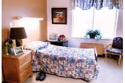 A single bed with a patterned bedspread in a well-lit room. Next to the bed is a wooden nightstand with a lamp, a framed photo, and a small flower arrangement. There is a window with floral curtains letting in natural light, a framed picture on the wall, a small table with a microwave, and a chair with a cushion near the window.