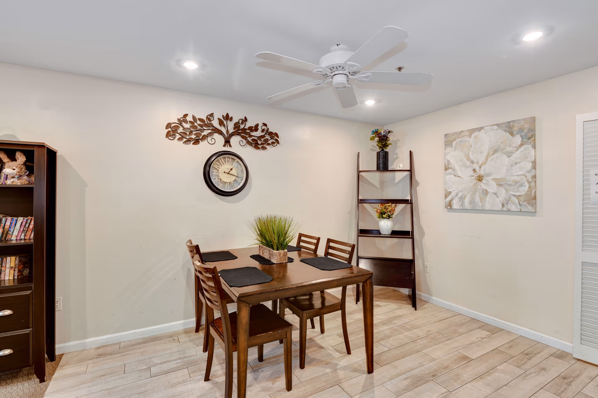 A dining area with a wooden table and four chairs, a decorative wall clock with a metal leaf design above it, a wooden shelf with vases and flowers, a large floral painting on the wall, and a ceiling fan with lights.