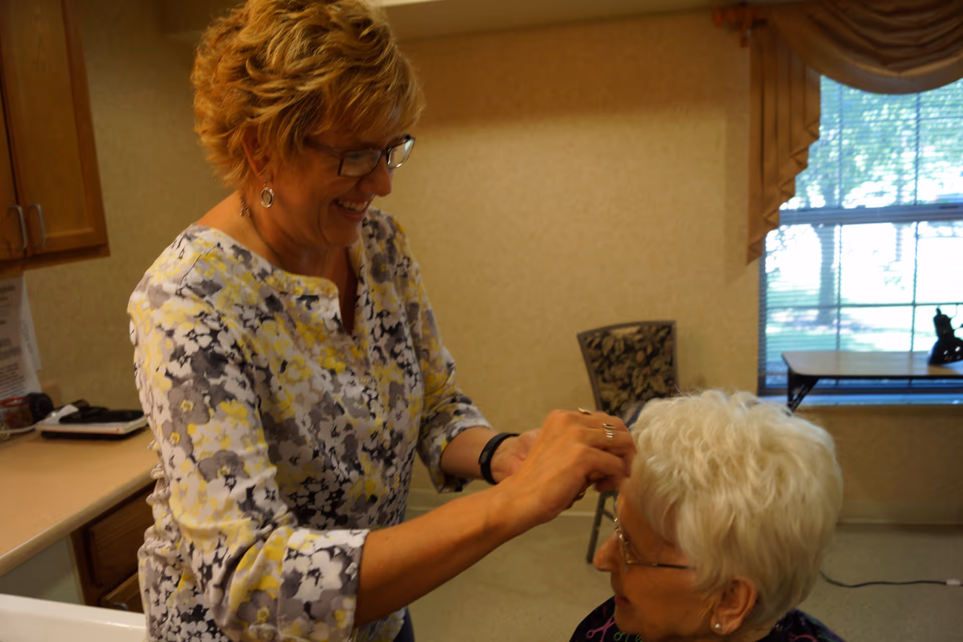A woman with short curly hair and glasses is smiling and gently touching the hair of an elderly woman with white hair and glasses in a cozy room with a window and a chair in the background.
