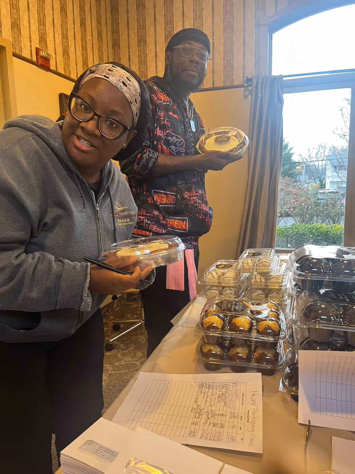 Two people standing indoors near a table with multiple clear plastic containers of baked goods. One person is wearing glasses, a headband, and a gray hoodie, holding a container and smiling at the camera. The other person is wearing glasses, a black head covering, and a patterned shirt, also holding a container. There is a window with curtains in the background letting in natural light.