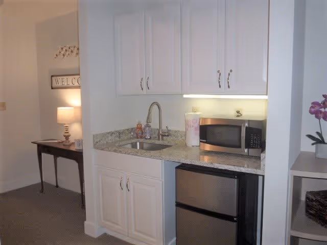 Small kitchenette area with white cabinets, a granite countertop, a stainless steel sink with a faucet, a microwave, a mini refrigerator, and a paper towel holder. To the left, there is a small table with a lamp and a wall decoration that says 'WELCOME'.