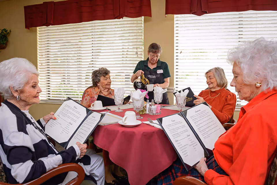 A server pours coffee for four elderly women seated around a table in a retirement center dining room as they hold menus.