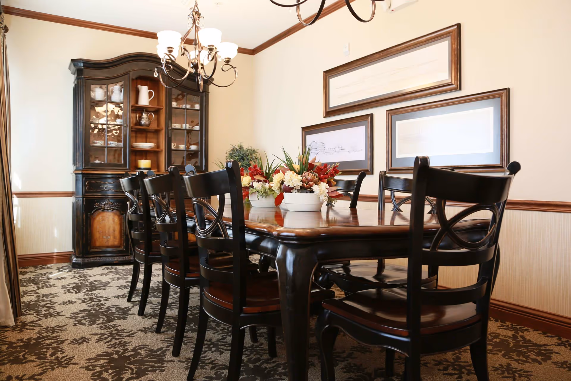 A dining room with a long wooden table surrounded by black wooden chairs. The table has floral centerpieces with red, white, and green flowers. On the wall behind the table are three framed artworks. To the left, there is a dark wooden china cabinet displaying white dishes and pitchers. A chandelier with multiple lights hangs above the table. The room has beige walls with wooden trim and patterned carpet flooring.