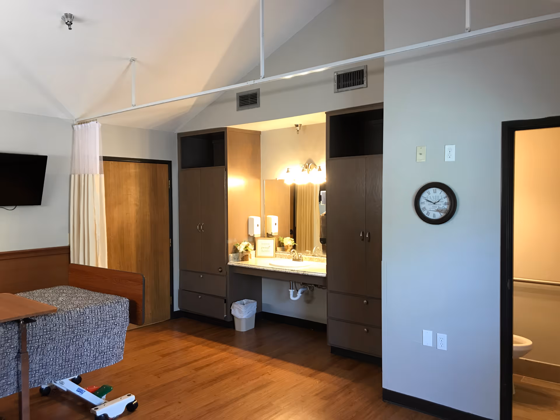 A senior living facility room with a hospital-style bed covered in a patterned blanket, a wall-mounted TV, a wooden door, a sink area with cabinets and a mirror, a clock on the wall, and a partially visible bathroom with a toilet.