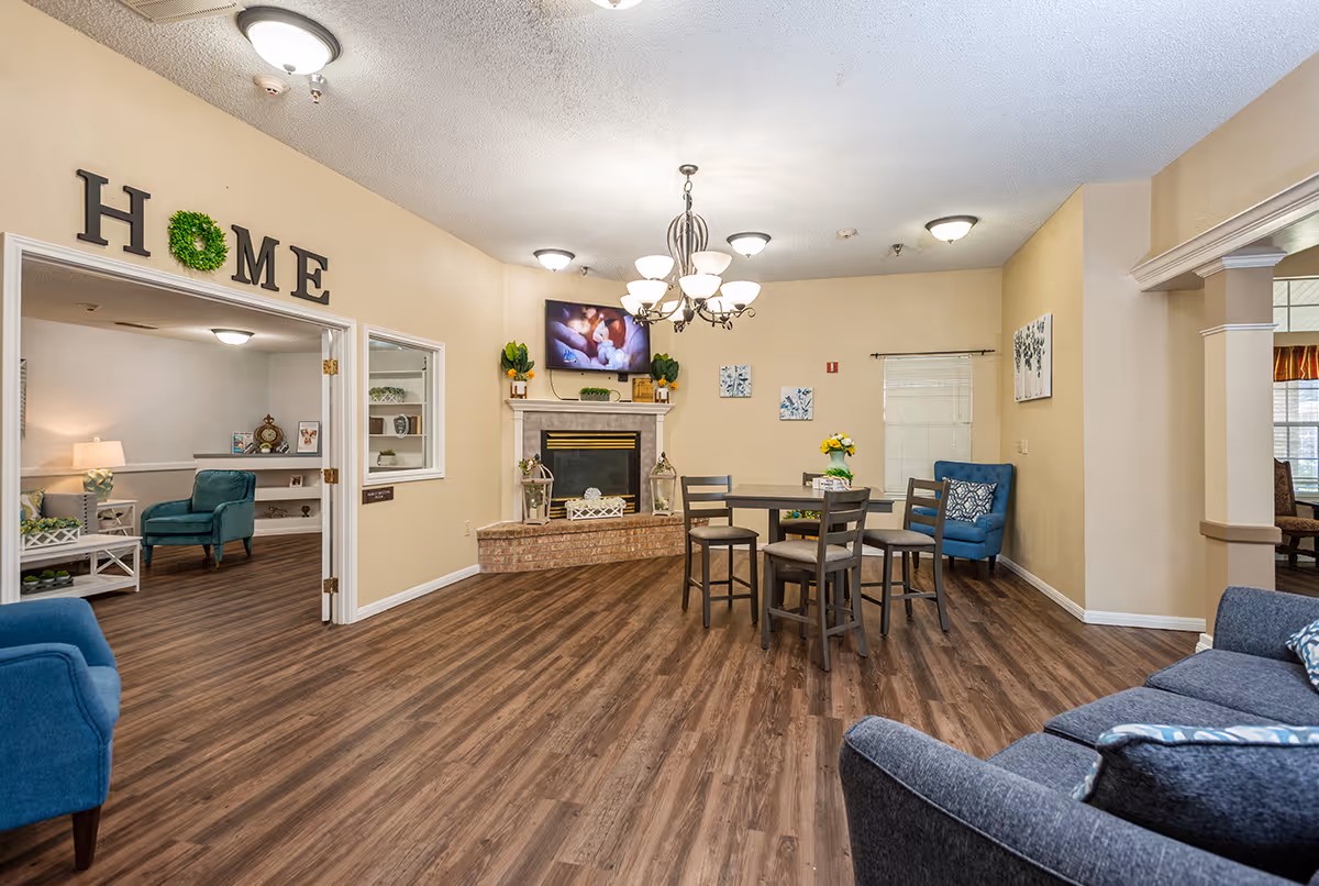 Community living room with a fireplace and TV, seating areas, and a small dining table under a chandelier.