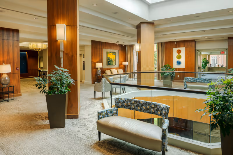 Spacious, well-lit lounge area with seating, potted plants, wooden columns, and a glass-railed atrium.