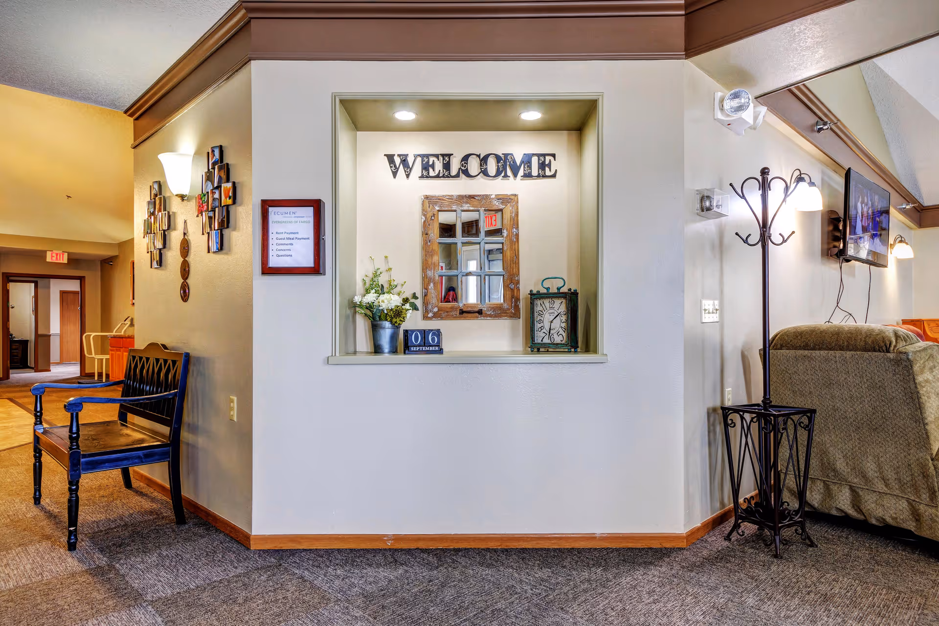 Interior view of a senior living facility lobby area with a decorative niche featuring a 'WELCOME' sign, a mirror, a clock, and a flower pot. To the left, there is a black wooden bench and a wall with photo frames. To the right, there is a coat rack, a wall-mounted TV, and a beige armchair. The space is well-lit with wall sconces and recessed lighting.