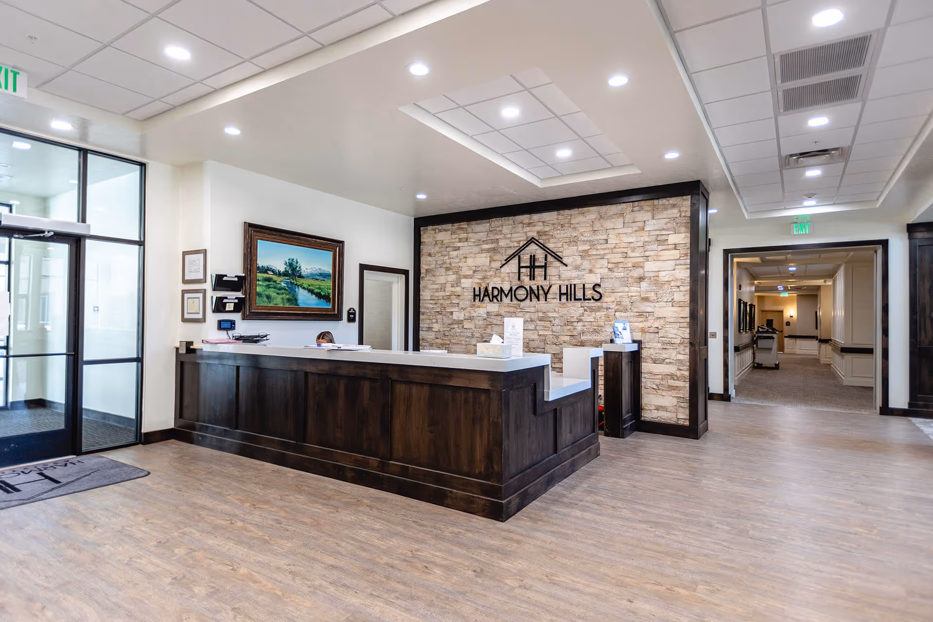 Bright reception lobby with a dark wood front desk, stone accent wall reading "Harmony Hills", glass entry doors and a hallway to the right.