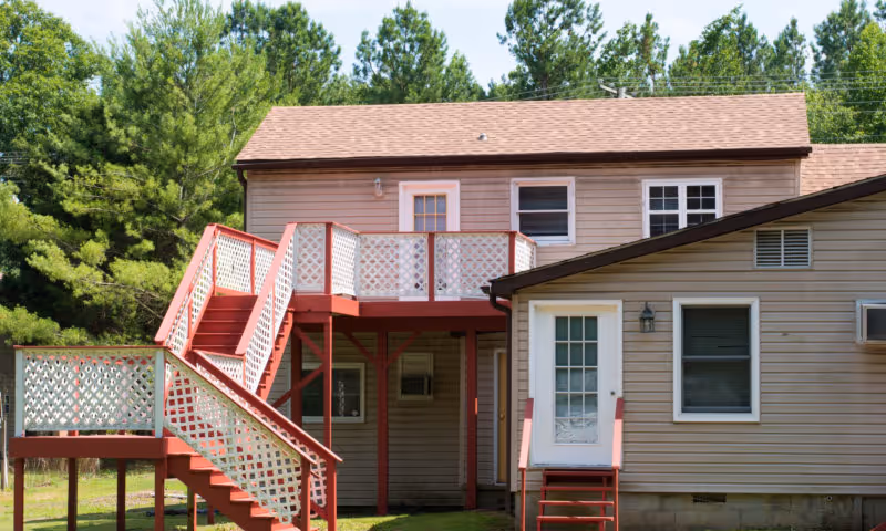 Exterior view of a two-story building with beige siding and a brown shingled roof. The building features a red wooden staircase and balcony with white lattice railings. There are several windows and a white door with glass panels leading to the balcony. The background includes green trees under a clear sky.