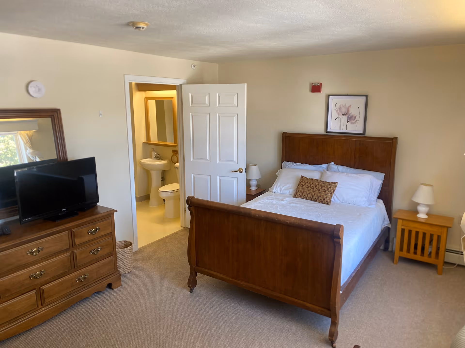 A senior living bedroom with a wooden bed frame, white bedding, and a decorative pillow. There are two bedside tables with lamps on either side of the bed. A dresser with a flat-screen TV and a large mirror is positioned against one wall. The door to an adjoining bathroom is open, showing a pedestal sink and toilet inside. The room has beige walls and carpeted flooring.