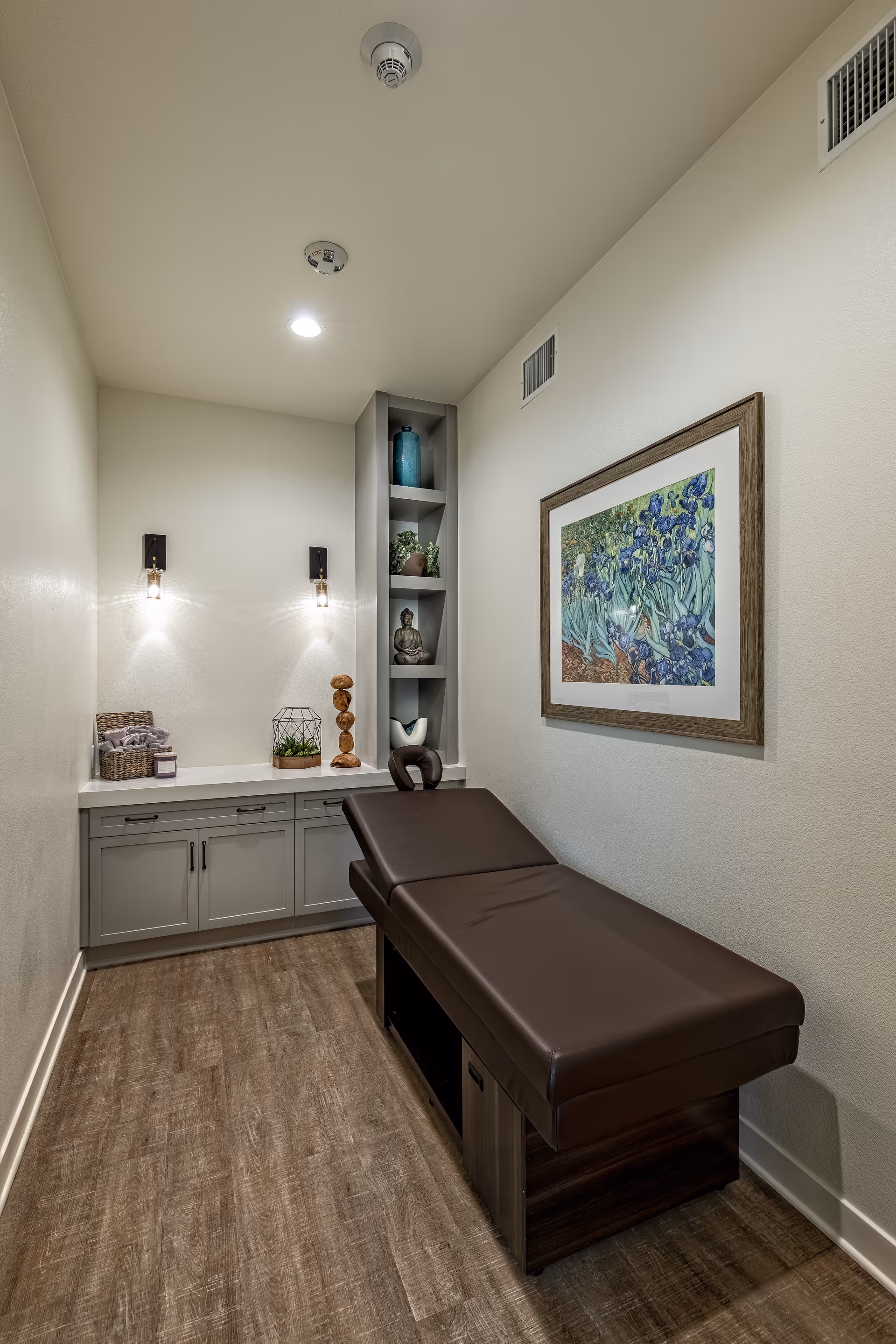 A small, well-lit room with a brown cushioned massage or therapy table, light gray cabinetry with decorative items on the countertop, built-in shelving with plants and sculptures, and a framed floral painting on the wall.