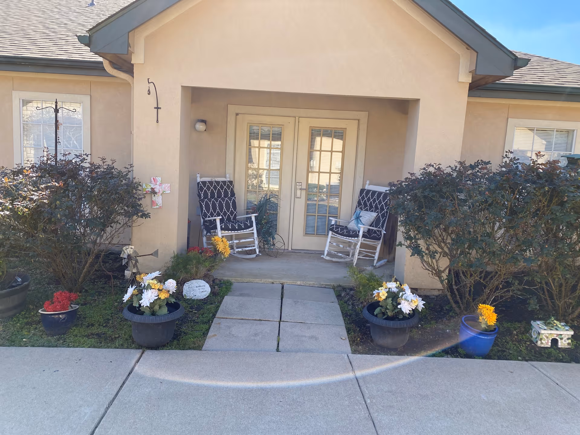 Entrance to a building with two white rocking chairs with black and white cushions on a small porch. There are potted flowers and bushes on either side of the concrete walkway leading to the door. The building has beige walls and a double glass door with blinds.