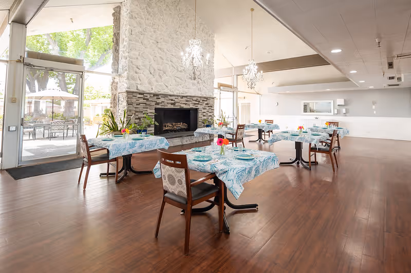 Dining room with tables set with blue patterned tablecloths, wooden chairs, a large stone fireplace, chandeliers, and glass doors leading to an outdoor patio.
