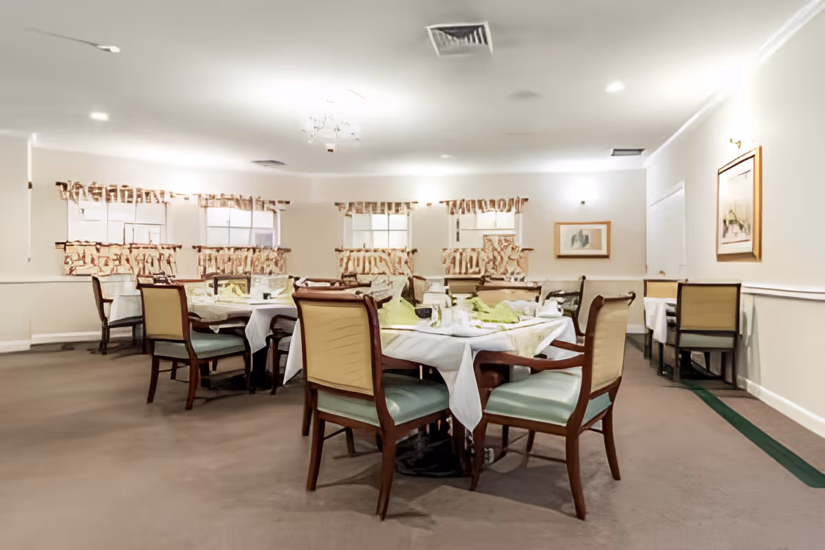 Communal dining room with round tables set with white tablecloths and chairs beneath curtained windows.