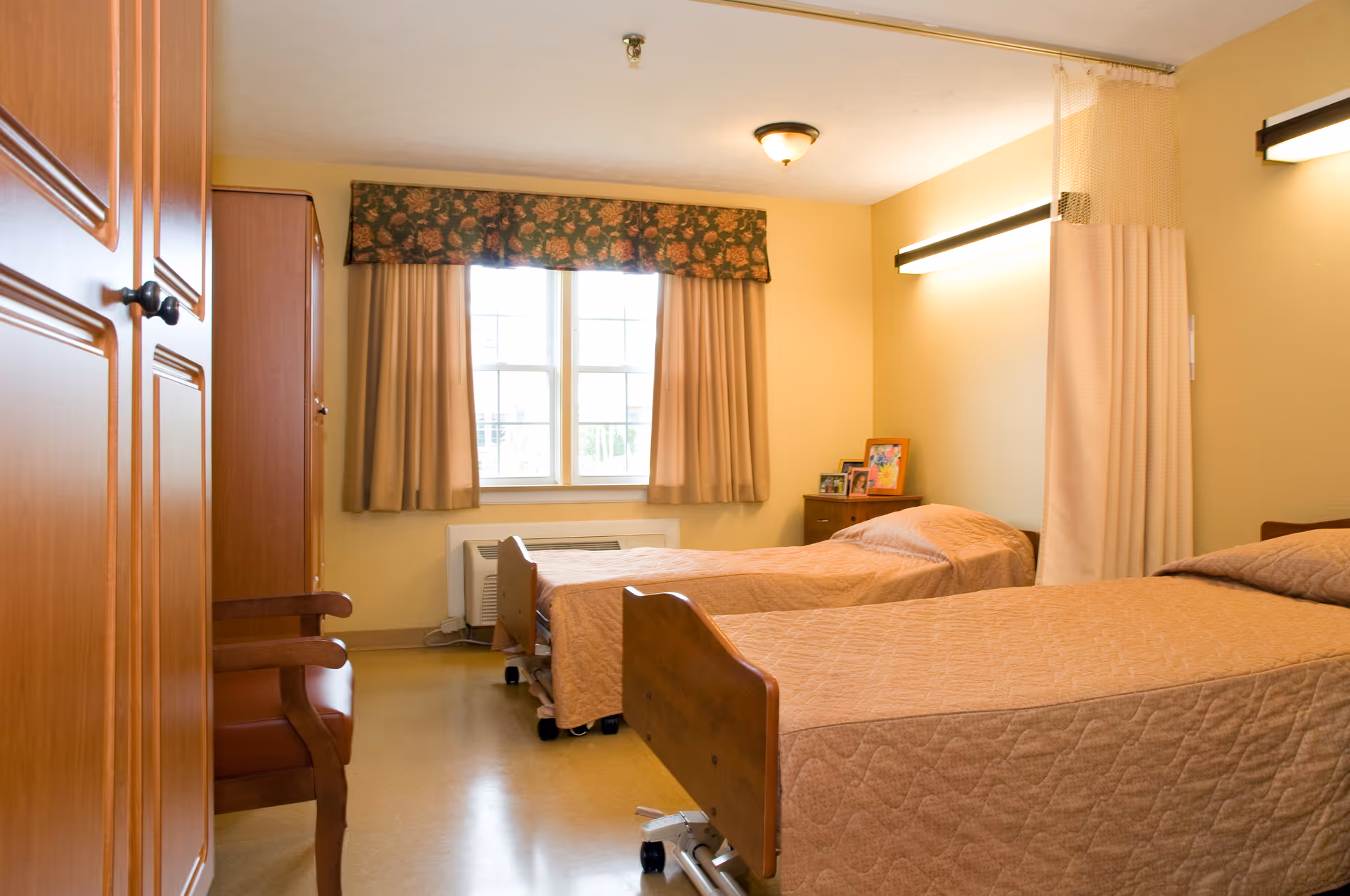 Two single beds with beige quilted bedspreads in a warm, well-lit senior care bedroom with a window, wardrobe and bedside table.