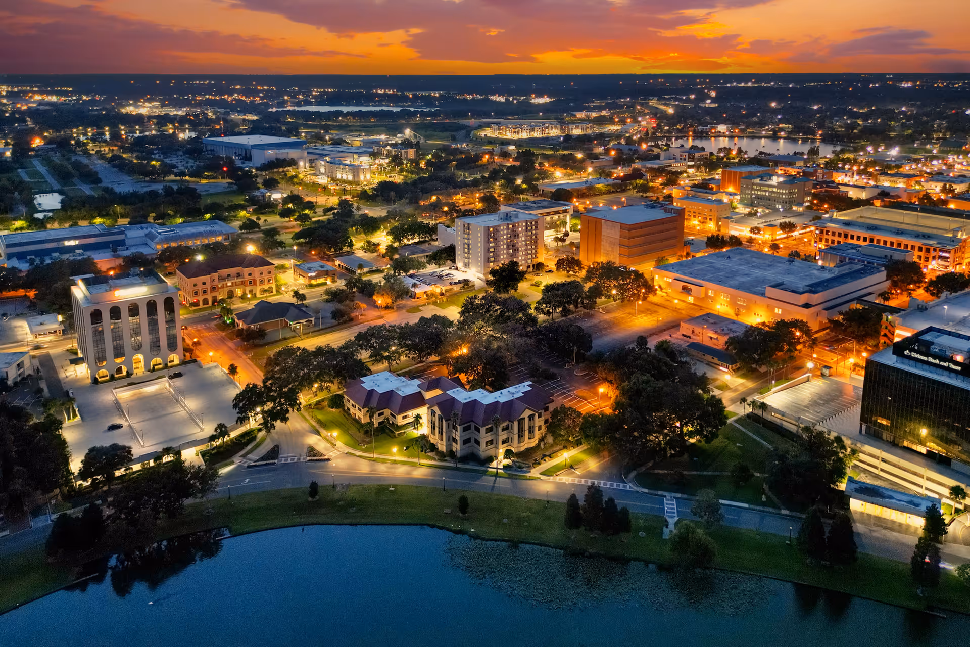 Aerial view of a cityscape at sunset with buildings, streets, trees, and a body of water in the foreground. The sky is vibrant with orange and purple hues as city lights begin to illuminate the area.