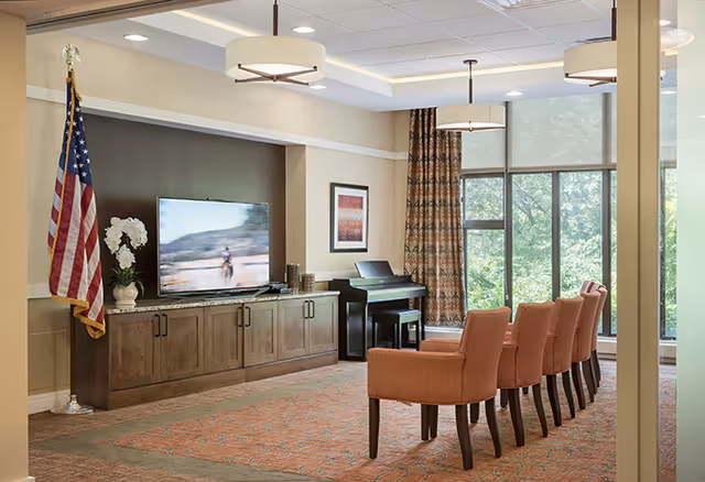 A well-lit common area with a row of five orange upholstered chairs facing a large flat-screen TV mounted on a wooden cabinet. To the left of the TV is a potted white orchid and an American flag on a stand. A piano with a bench is positioned against the wall under a framed picture. Large windows with patterned curtains allow natural light to fill the room.
