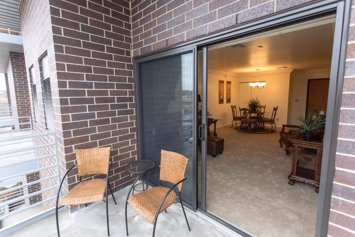 Small balcony with two wicker chairs and a side table in front of a sliding glass door opening into a furnished living and dining room.