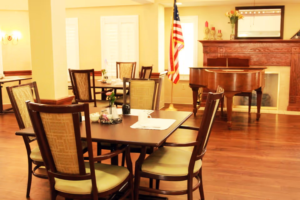 Dining area with wooden tables and chairs, a grand piano by a fireplace, and an American flag.