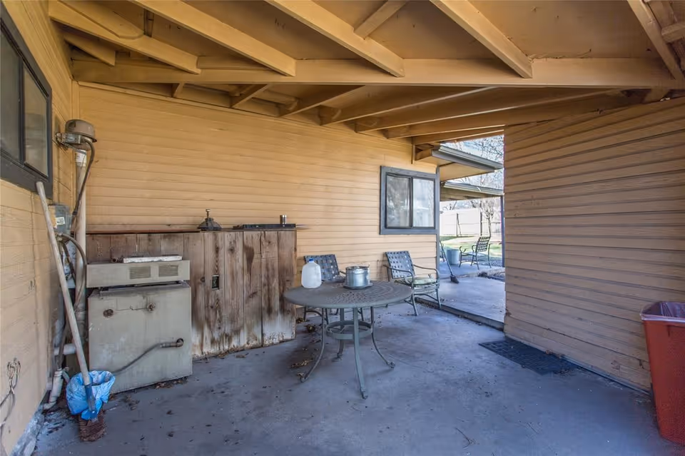 Covered outdoor patio area with a round metal table and chairs, a wooden storage cabinet, and various utility items against a beige siding wall.