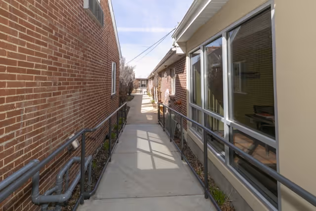 A narrow outdoor walkway between two buildings with brick and beige walls, metal handrails on both sides, and a clear sky above.