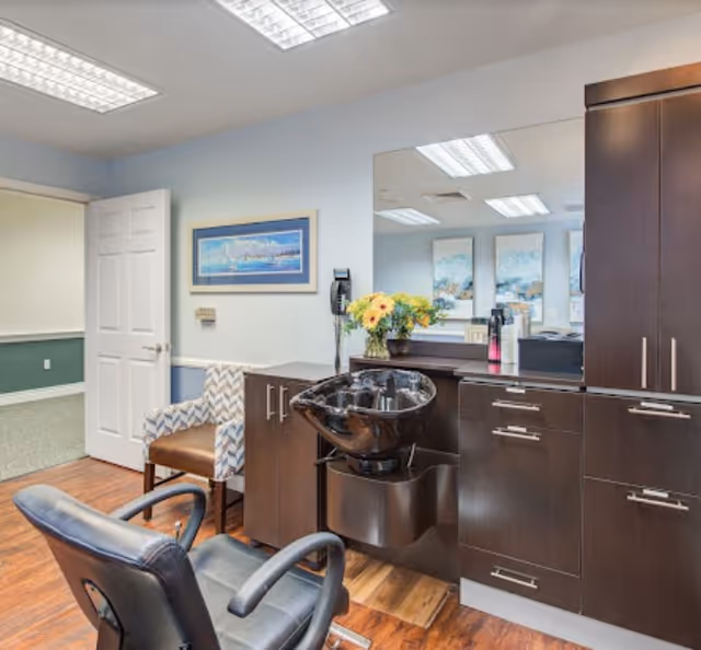 Interior of a salon area in a senior living facility with a black salon chair and a black hair washing sink attached to dark wooden cabinetry. A patterned armchair is positioned near an open door leading to another room. A large mirror is mounted above the cabinetry, reflecting parts of the room. A framed picture hangs on the light blue wall, and a vase with yellow flowers is placed on the counter.