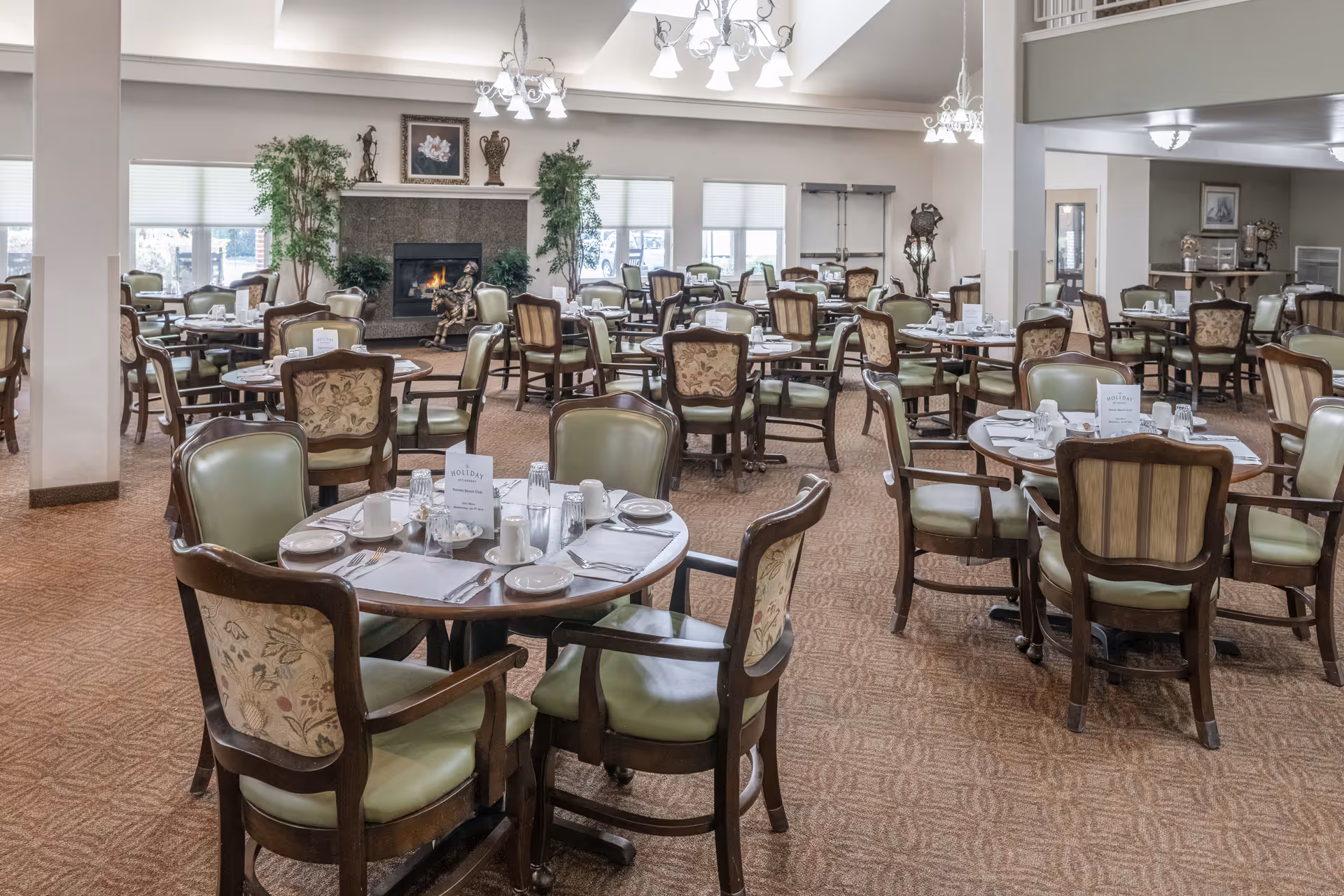 A spacious dining room with multiple round tables set with plates, cups, and silverware. The chairs have wooden frames with green cushioned seats and floral-patterned backs. In the background, there is a fireplace with decorative items on the mantel and large windows letting in natural light. The room has a warm, inviting atmosphere with carpeted floors and chandeliers hanging from the ceiling.