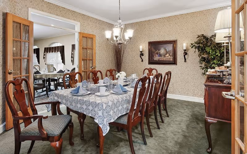 A formal dining room with a long wooden table covered with a patterned tablecloth, set with plates, cups, and folded napkins. The room features wooden chairs with cushioned seats, a chandelier hanging above the table, a framed painting on the wall, two wall-mounted candle holders, and a sideboard with decorative items and a lamp. French doors open to an adjacent room with additional seating and windows with valances.