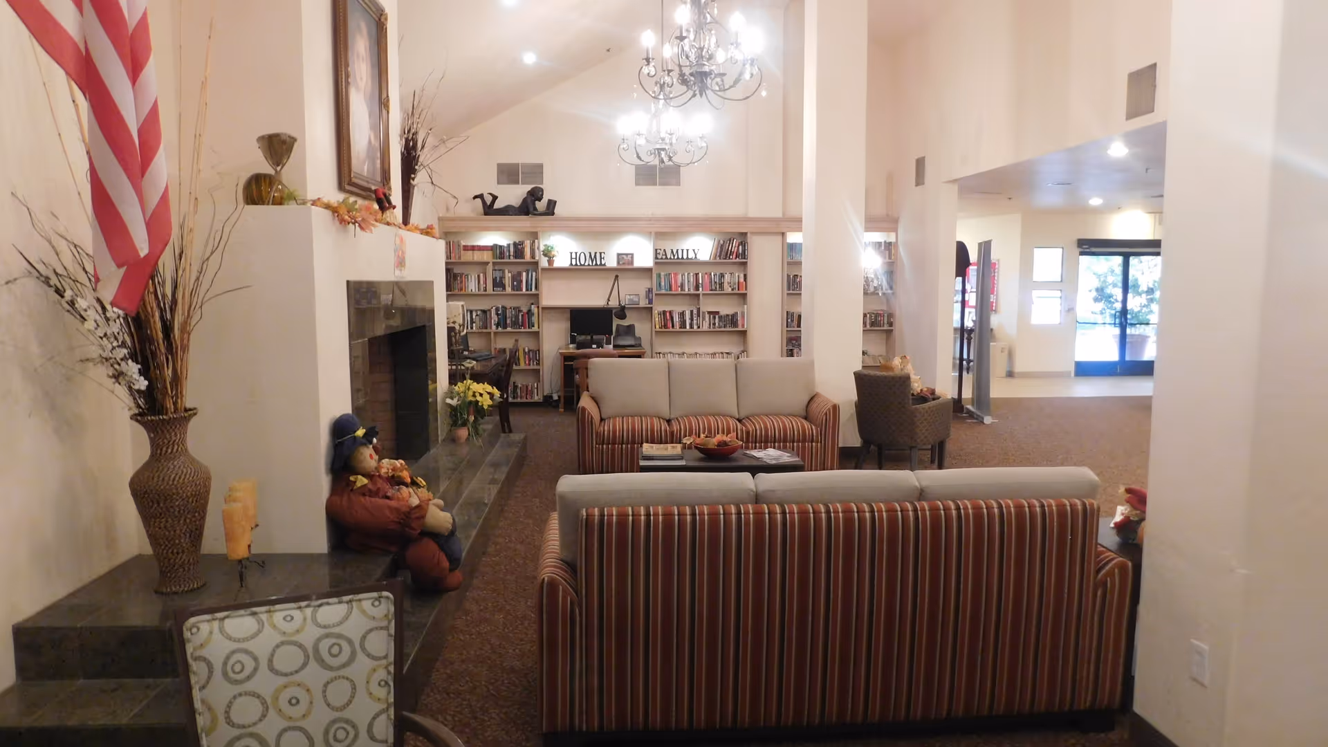 Community living room with striped sofas arranged around a fireplace, bookshelves, chandeliers, and an entrance in the background.