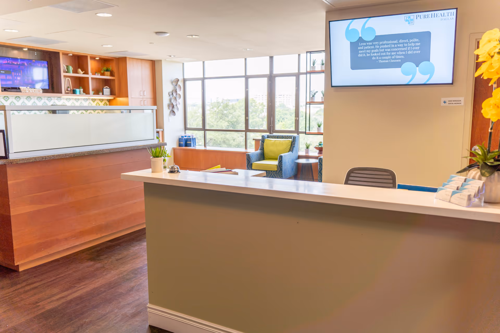 Reception area of PureHealth Transitional Care at THR Arlington with a front desk, a chair with a green cushion near large windows, a wall-mounted TV screen displaying a testimonial, and decorative plants on the counter and shelves.
