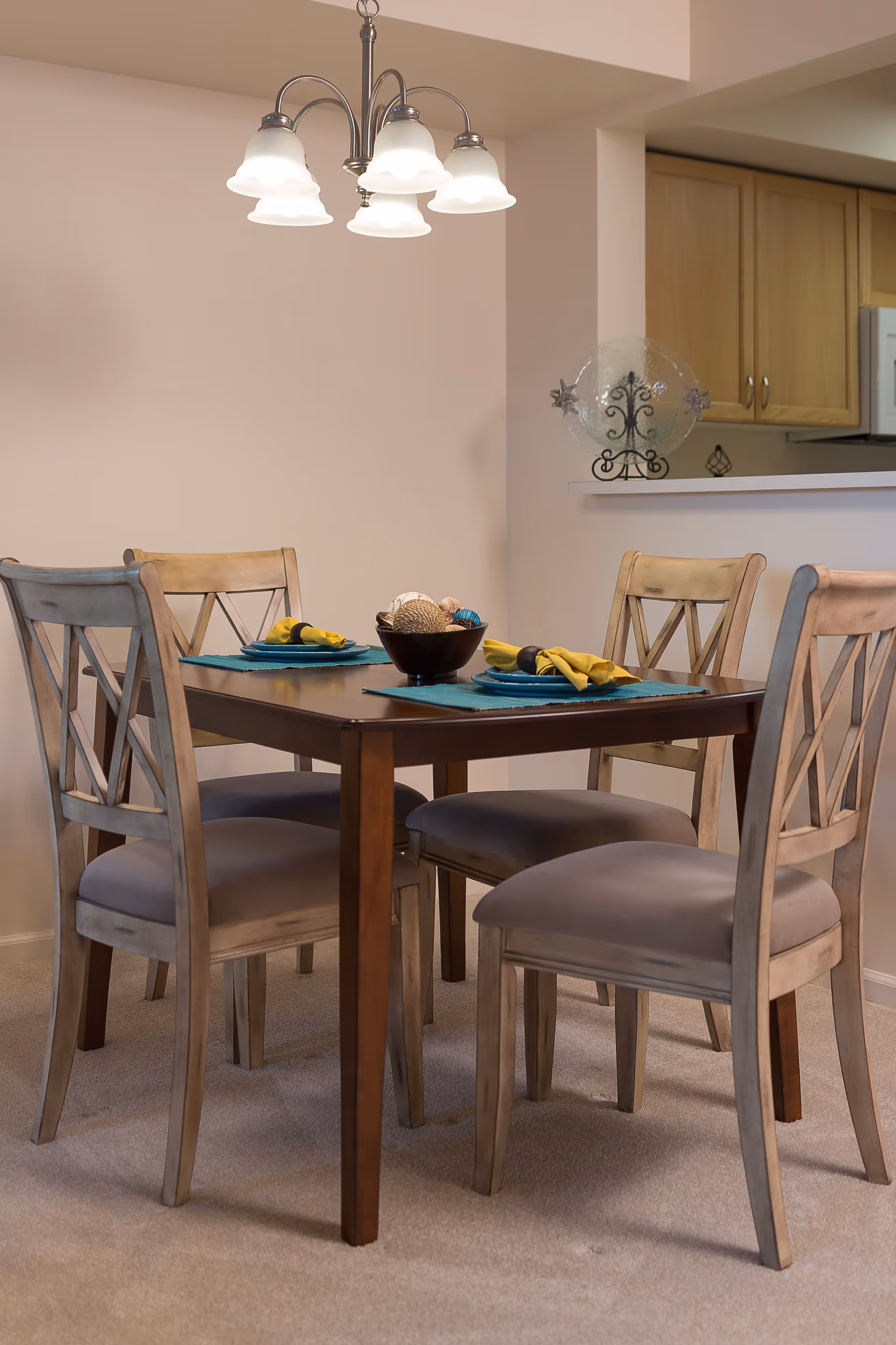 A dining area with a wooden table set for four, featuring four cushioned chairs with wooden frames. The table is decorated with blue placemats, yellow napkins, and a centerpiece bowl with decorative balls. Above the table hangs a chandelier with five frosted glass shades. In the background, there is a partial view of a kitchen with wooden cabinets and a countertop.