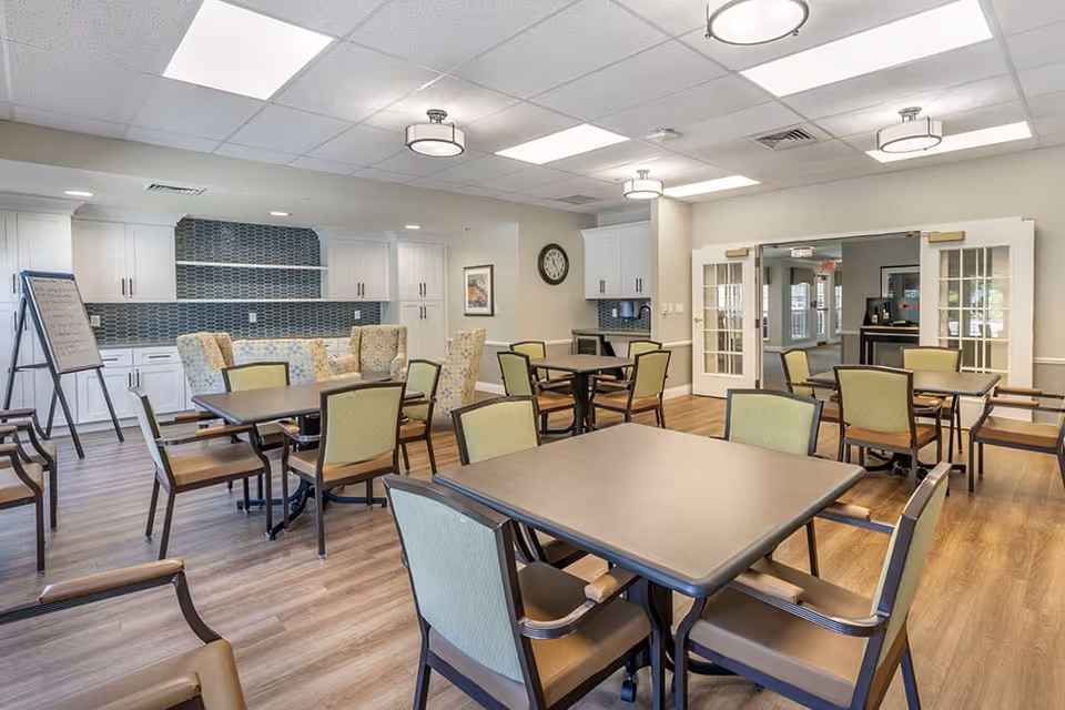 A bright and spacious dining area in a senior living facility with multiple tables and chairs arranged neatly. The room features wood flooring, white cabinetry with a blue tile backsplash, a clock on the wall, and a whiteboard on an easel. There are also comfortable armchairs and double glass doors leading to another room.