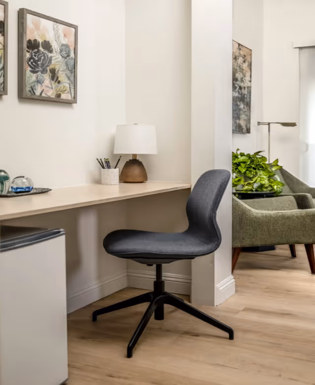 A small workspace with a modern black swivel chair in front of a light wooden desk. On the desk, there is a lamp with a white shade, a container holding pencils, and decorative items. The wall above the desk features framed floral artwork. In the background, there is a green armchair, a floor lamp, and a leafy green plant near a window with a white blind.