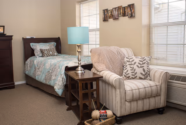 A furnished bedroom with a twin bed, nightstand and lamp, striped armchair, dresser, and two windows.