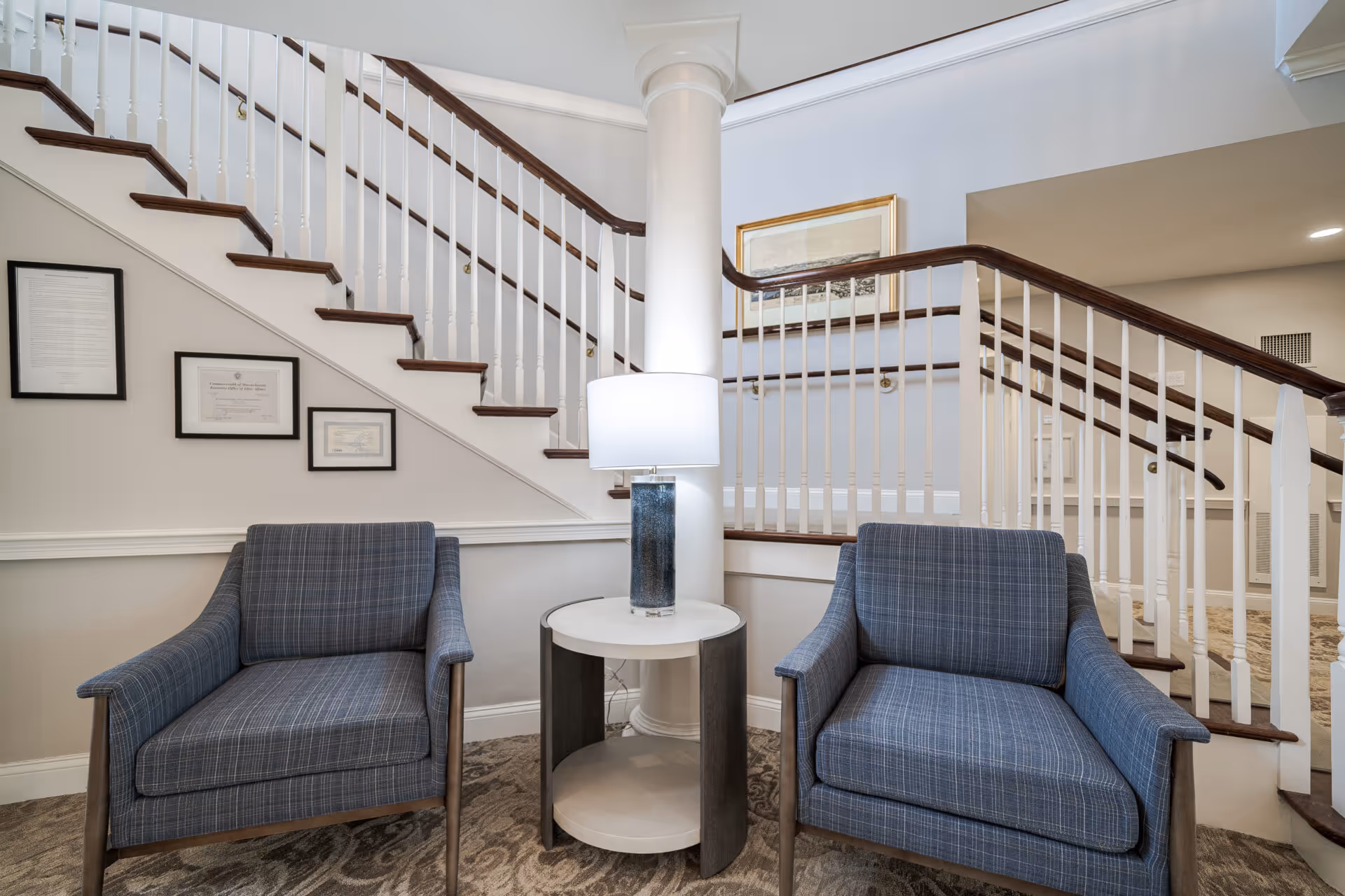 Two blue upholstered armchairs flanking a round side table with a lamp in a seating area at the base of an interior staircase.