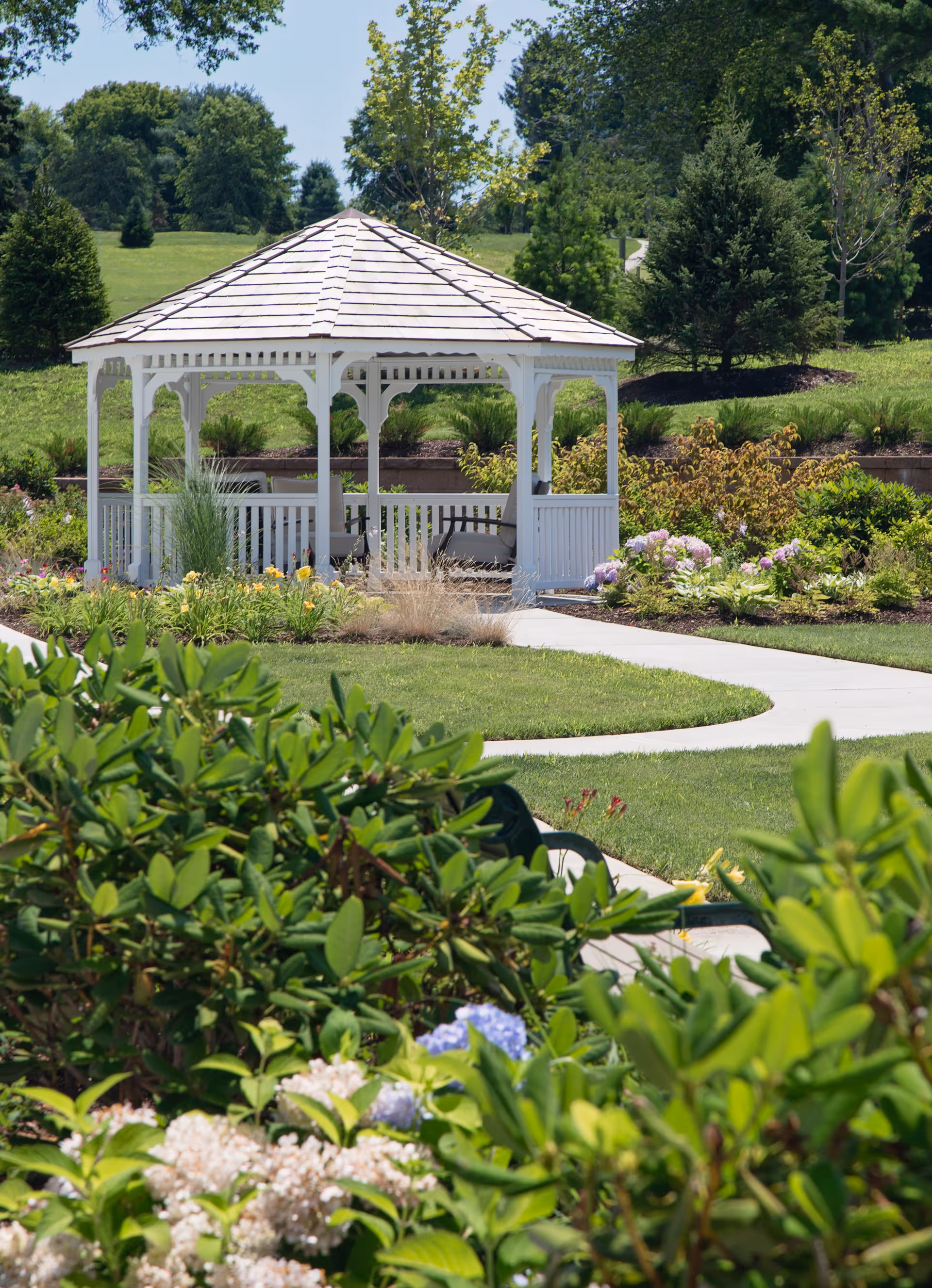 White wooden gazebo surrounded by landscaped gardens, flowering shrubs, and a winding paved path.
