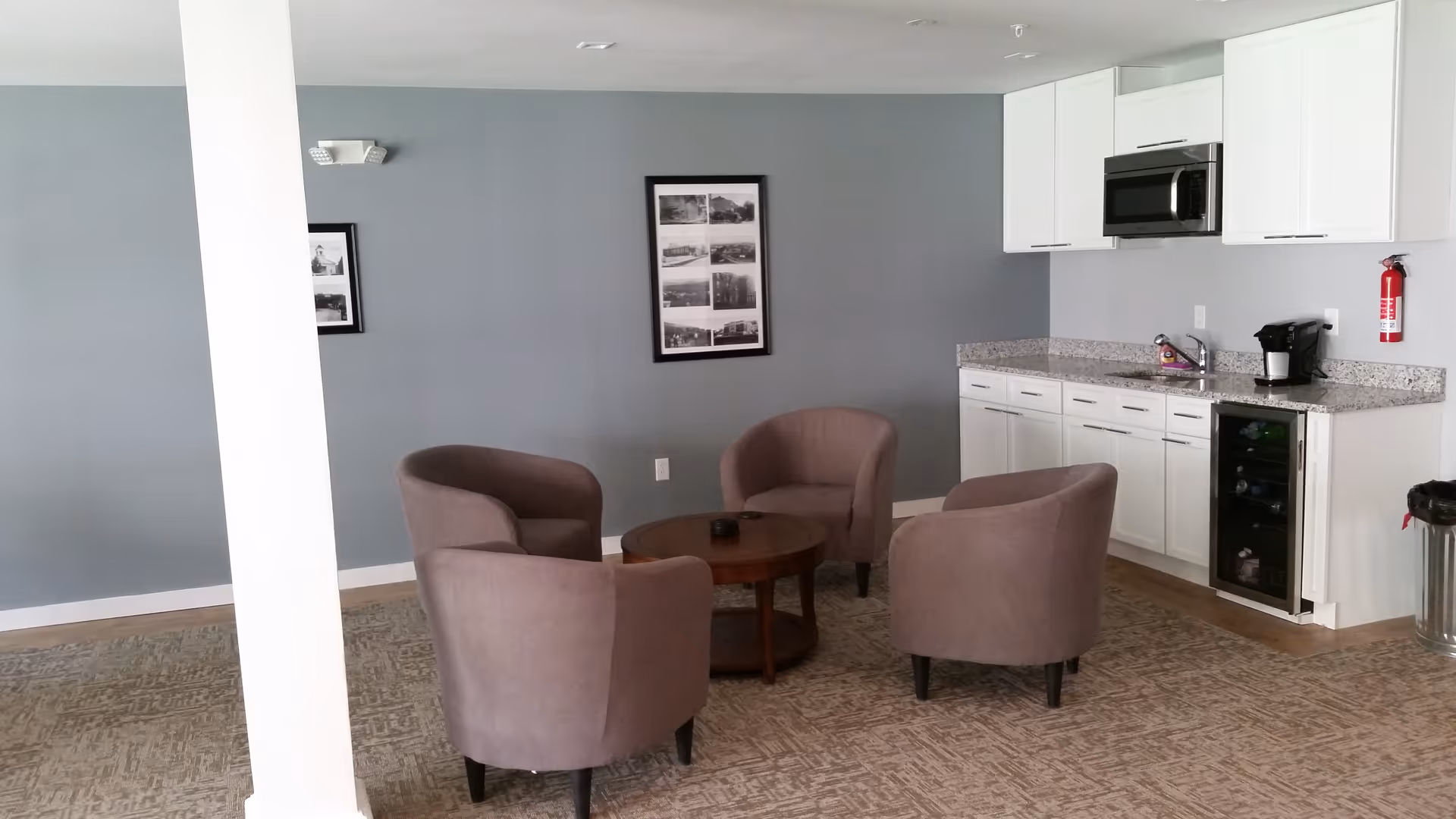 A common area with four gray upholstered armchairs arranged around a round wooden coffee table. The room has gray walls with framed black and white pictures, a kitchenette with white cabinets, granite countertops, a microwave, a coffee maker, and a small wine cooler. There is a fire extinguisher mounted on the wall and a trash can nearby.
