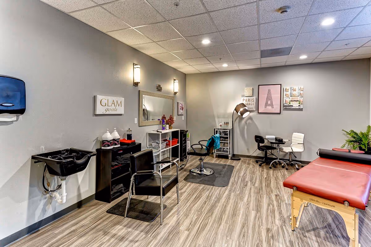 Interior view of a beauty and wellness room in an assisted living facility. The room features a black hair washing sink with a chair, a styling chair in front of a large mirror, shelves with hair products and towels, a manicure table with two chairs, a floor lamp, and a massage table with a red cushion. The walls are decorated with framed pictures and a sign that reads 'GLAM goals'. The floor has wood-patterned vinyl flooring and the ceiling has recessed lighting.