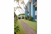 A paved walkway runs alongside a blue and white multi-story building with balconies. The path is bordered by green grass and tropical plants, with palm trees visible in the background under a clear sky.