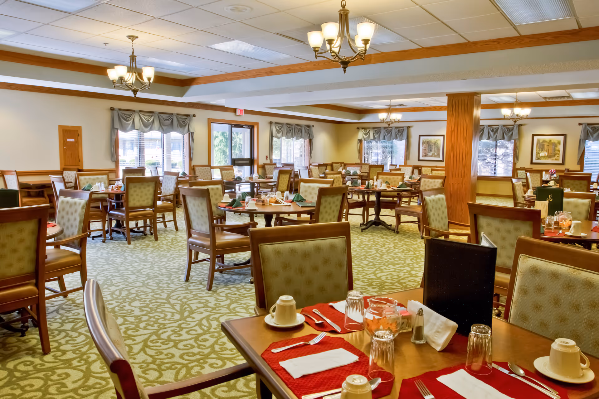 A spacious dining room with multiple wooden tables and cushioned chairs arranged neatly. Each table is set with cups, glasses, napkins, and utensils. The room has large windows with curtains, framed artwork on the walls, and ceiling lights with chandeliers providing warm lighting. The carpet has a green and beige pattern, and the overall ambiance is cozy and inviting.