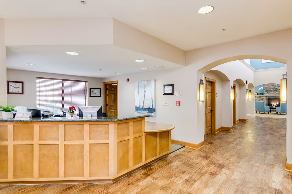 Reception area with a wooden front desk and granite countertop, two computer monitors, a potted plant, and a small red flower arrangement. Behind the desk is a window with blinds, a clock on the wall, and framed certificates. To the right, a hallway with wooden doors and wall-mounted lights leads to a seating area with blue chairs and a stone fireplace.