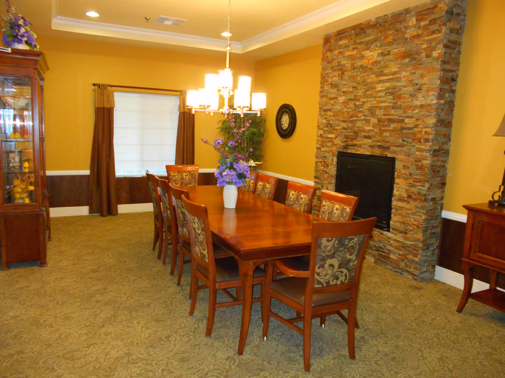 A dining room with a long wooden table surrounded by eight chairs with patterned upholstery. A vase with purple flowers is placed in the center of the table. The room has yellow walls, a stone fireplace, a clock on the wall, a window with brown curtains, and a wooden cabinet displaying decorative items.