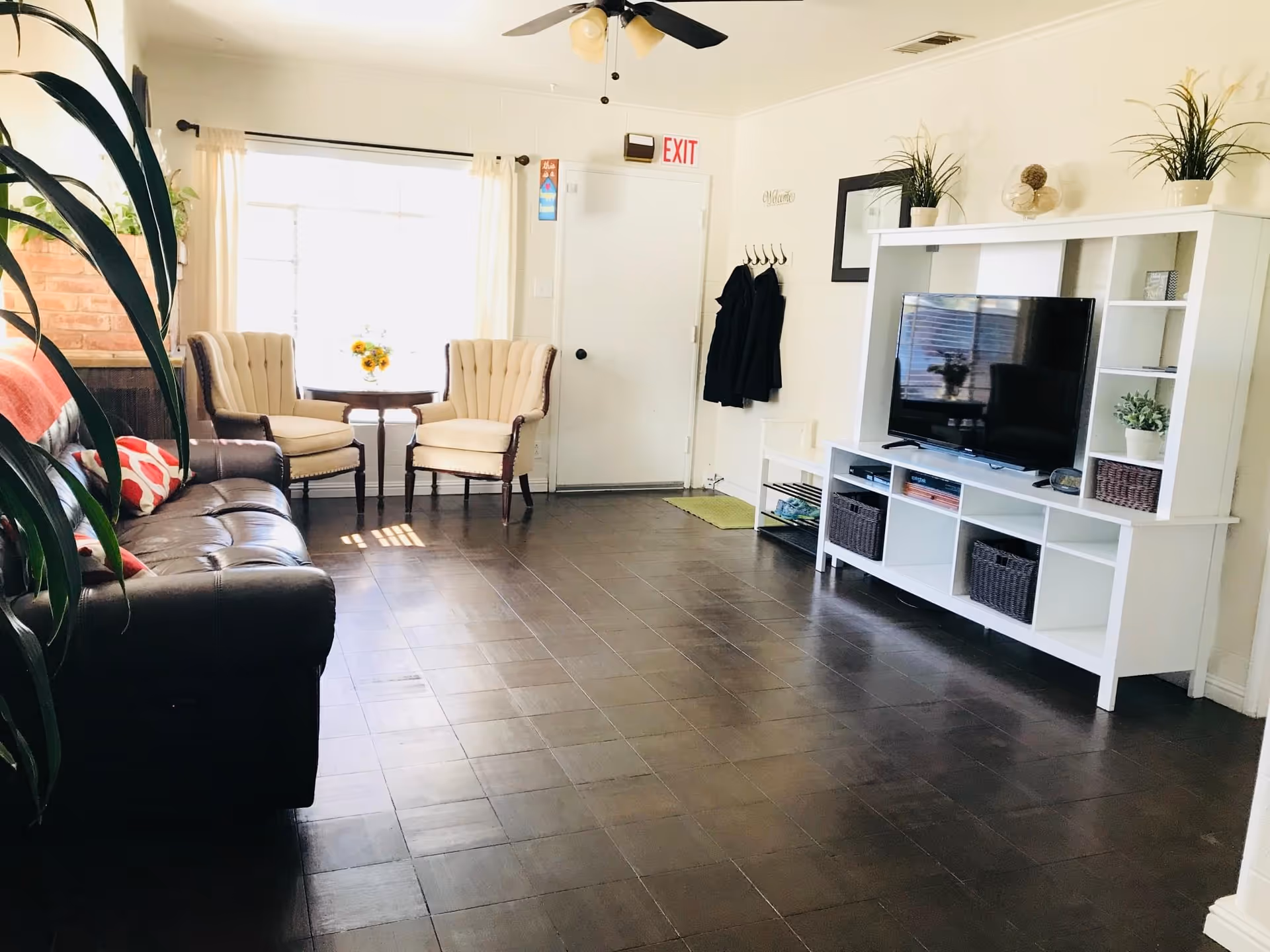 A bright living room with a dark brown leather sofa on the left, two beige armchairs with a small round table between them near a window with white curtains. On the right side, there is a white entertainment unit with a flat-screen TV, decorative plants, and storage baskets. The floor is dark wood, and there is a ceiling fan with lights above. A coat rack with black coats hangs near a white door with an exit sign above it.