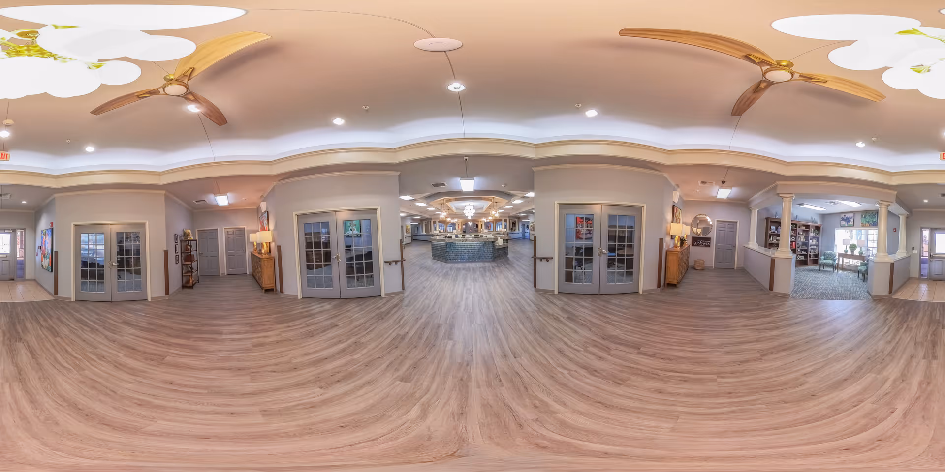 Panoramic interior view of a senior living facility lobby with wood floors, a central reception desk, seating areas, and double doors.