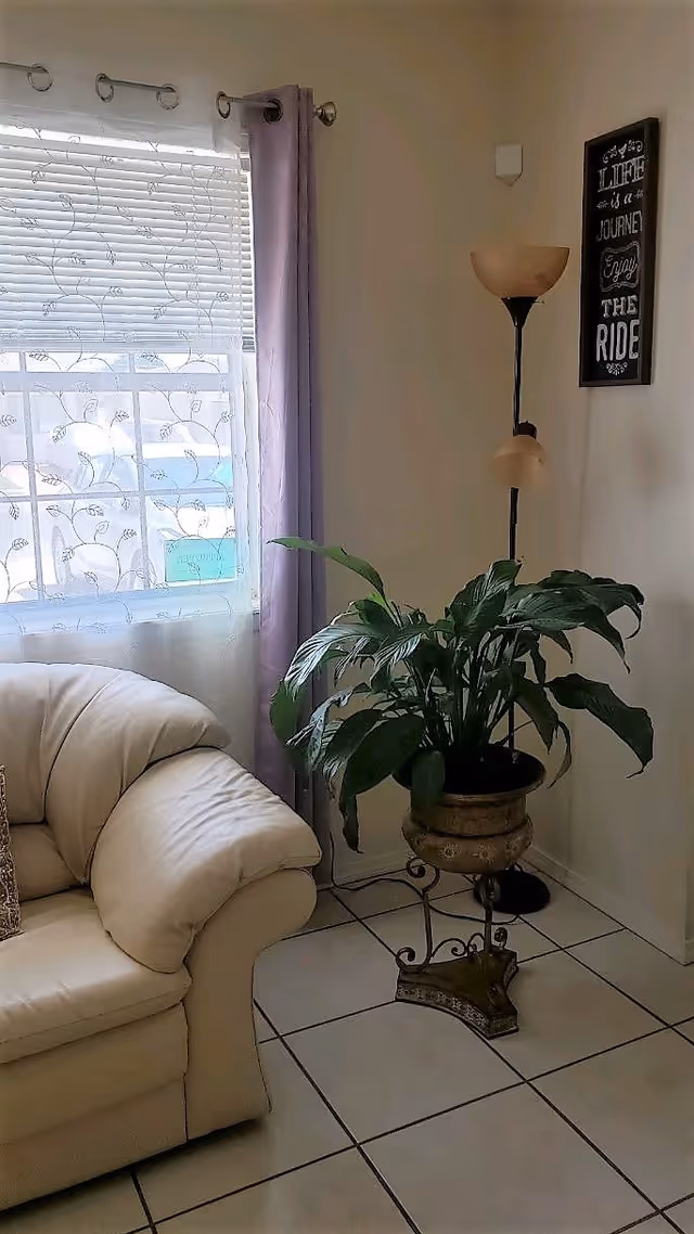 A cozy corner of a living room featuring a cream-colored leather sofa, a window with sheer floral curtains and purple drapes, a tall floor lamp with two beige lampshades, and a large green potted plant on an ornate metal stand. A decorative wall sign with the words 'Life is a journey, enjoy the ride' is hung on the wall.